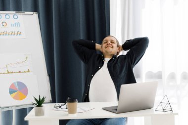 Günlük kıyafetler giymiş bir adam ofiste dizüstü bilgisayarla çalışır ve olumlu duygularını ifade eder. Adam ücra bir yerde çalışıyor.