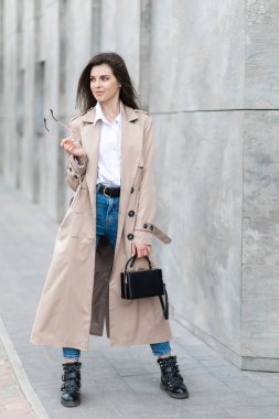 Yağmurluk ve kot pantolonlu genç bir kız elinde el çantasıyla şehrin etrafında dolanıyor..