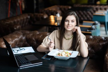 Genç bayan kafede dizüstü bilgisayarla oturmuş salata yiyor.
