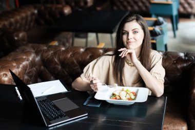 Genç bayan kafede dizüstü bilgisayarla oturmuş salata yiyor.