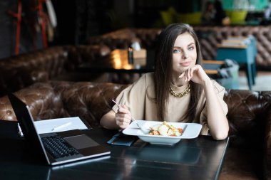 Genç bayan kafede dizüstü bilgisayarla oturmuş salata yiyor.