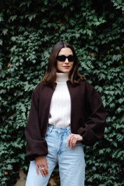 Şık, genç bir kadın, sarmaşık kaplı bir duvarın önünde güven içinde poz veriyor, son moda sonbahar kıyafetlerini sergiliyor.