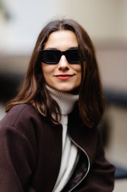 Kahverengi ceket giyen şık bir kadın, balıkçı yaka kazak ve kot pantolon, şehir ortamında güven içinde poz veriyor.