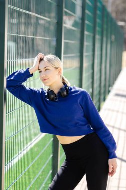 Kulaklık takan sporcu kadın antrenmandan sonra spor sahasının çitlerine yaslanıyor.