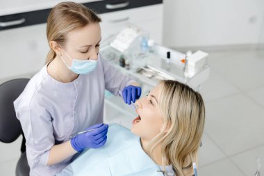 Diş hekiminde maske takan ve eldiven takan bir diş hekimi hasta dişlerini diş aletleriyle muayene ediyor.