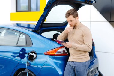 Genç bir adam bir istasyonda durup tablet tutarken elektrikli bir arabayı şarj ediyor..