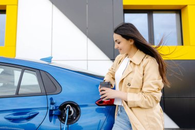 Küçük bir kamu istasyonundan şarj olan mutlu genç bir kadın, elektrikli arabanın yanında duruyor, kahve içiyor..