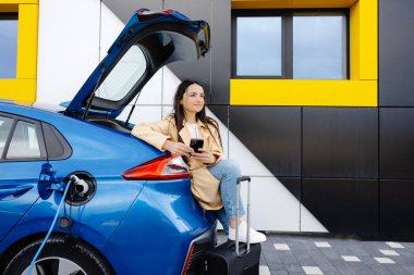 Genç bir kadın, küçük bir kamu istasyonundan şarj edilen otomobil aküsünü doldurmak için bekliyor ve otomobili şarj ederken akıllı telefon kullanıyor.