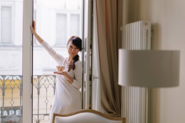 Adult woman enjoying a relaxing morning holding a cup of coffee or tea on a Parisian apartment balcony, smiling at the camera