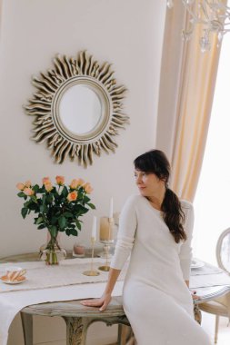 Woman in elegant white dress sitting on a stylish dining table, surrounded by beautiful light and vintage decor. Feeling cozy