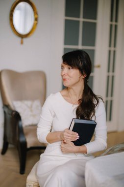 Woman holding a black book, looking at camera and smiling. Concept of knowledge, learning, and literature indoors