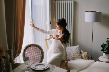 Zarif bir evde, pencerenin kenarında bir fincan çay tutan, dışarı bakan, sakin bir sabah anının tadını çıkaran bir kadın.