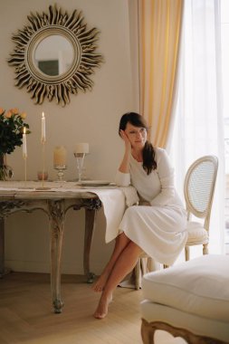 Yemek masasında çıplak ayakla oturan bir kadın dalgın dalgın dalgın dalgın dalgın dalgın dalgın dalgın dalgın dalgın dalgın bakıyor. Zarif iç tasarım sakin bir hava yaratıyor.