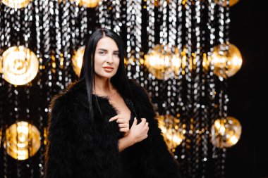 Young beautiful smiling brunette female in trendy evening fur coat. Carefree woman posing near shiny tinsel wall in studio. Fashionable model with bright makeup looks at camera.
