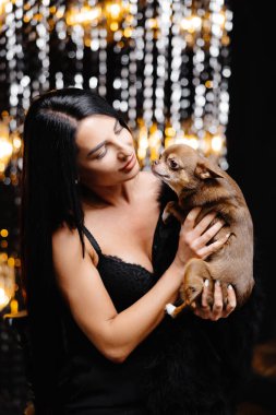 Young beautiful smiling brunette female in trendy evening clothes holding her litlle dog in her hands. Carefree woman posing near shiny tinsel wall in studio with dog. Fashionable model with bright makeup looks at camera.