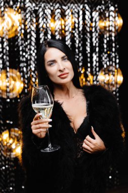 Young happy woman in trendy evening fur coat looking at camera and smiling with glass of champagne.
