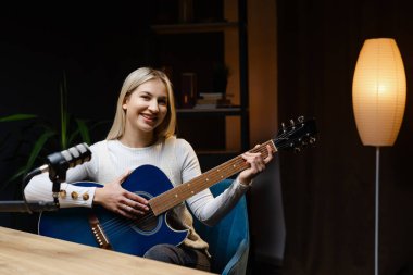 Müzisyen, profesyonel mikrofon ve mavi akustik gitar kullanarak ev stüdyosunda şarkı kaydediyor.