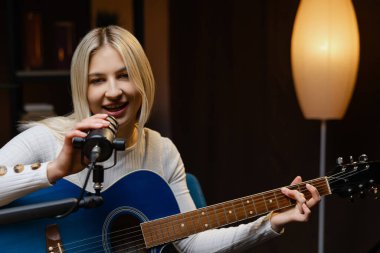 Müzisyen, profesyonel mikrofon ve mavi akustik gitar kullanarak ev stüdyosunda şarkı kaydediyor.