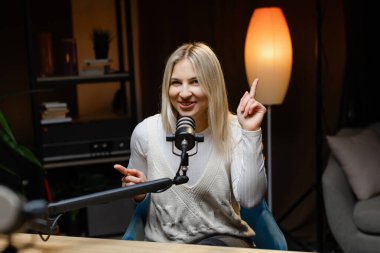 İçerik oluşturucu ses podcast 'ini mikrofon kullanarak kaydediyor ve stüdyoda masa başında otururken parmağıyla işaret ediyor.