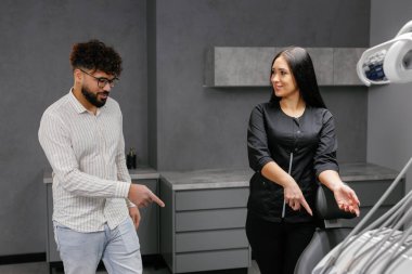 Kadın dişçi ya da asistan, dişçi koltuğunu işaret ederek, erkek hastaya modern bir klinikte muayeneye başlaması için selam veriyor.