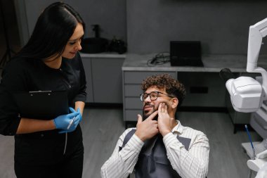 Modern bir dişçi muayenehanesindeki randevusu sırasında dişleri ağrıyan erkek hastaların yanında duran dişçi.