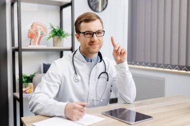 Gözlüklü ve beyaz üniformalı genç bir doktor masada oturuyor ve bir hastaya internetten danışıyor. Doktor parmağını kaldırarak ilgi istiyor..