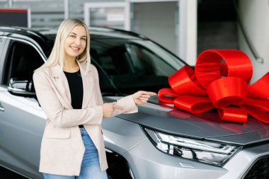 Satıcı kadın, galeride büyük kırmızı bir kurdeleyle süslenmiş gri bir arabayı işaret ediyor.