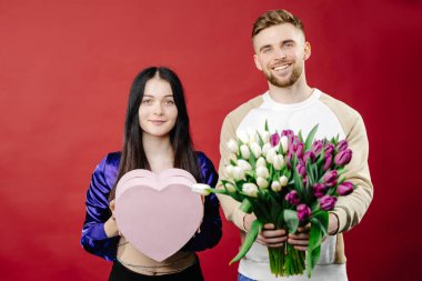 Gülümseyen sakallı adam güzel bir kadın için çiçek buketi ve hediye kutusu veriyor. Arka planda izole edilmiş mutlu romantik çiftin doğum günü portresi.