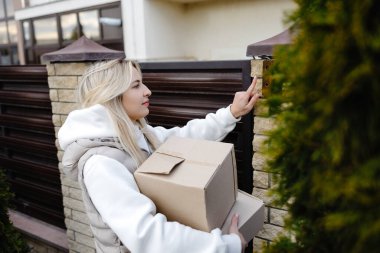 Bir evin zili çalarken elinde karton bir kutu tutan gülümseyen teslimatçı kadın teslimat servisini tamamlamaya hazır.