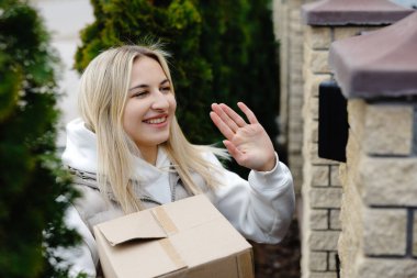 Teslimatçı kadın, elinde paketle evin girişinde gülümsüyor ve el sallıyor. Dışarıdaki dost canlısı müşteri hizmetlerini gösteriyor.