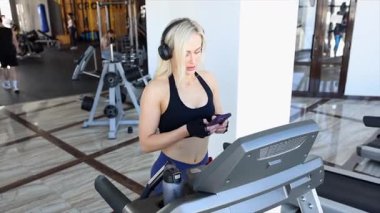 Spor salonunda akıllı telefon kullanan ve koşu bandında kulaklık takan atletik bir kadın.