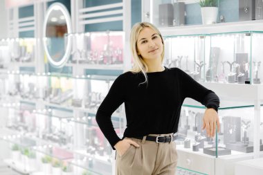 Bir kuyumcuda gülümseyen ve poz veren genç bir satıcı, eli cebinde ve bir vitrine yaslanmış.