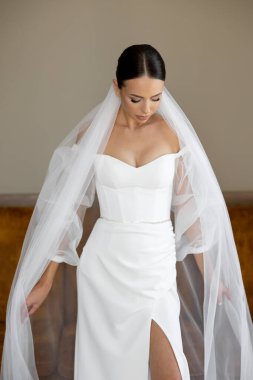 Gelin zarif, beyaz bir gelinlik ve duvak takmış, düşünceli bir şekilde aşağıya bakıyor, güzel ve romantik bir vücuda sahip.