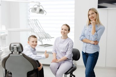 Diş hekimi bir anne ve oğluyla diş hijyeni hakkında konuşuyor, diş sağlığı kliniğinde oturuyor ve ayakta duruyor.