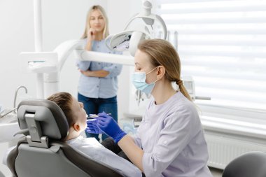 Annesi modern bir diş kliniğinde telefonla konuşurken genç bir çocuğun dişlerini muayene eden bir dişçi.