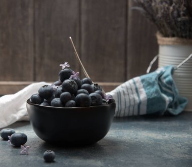 Üzerinde leylak çiçekleri olan taze yaban mersinleri mavi kıvrımlı bir tezgahın üzerinde, hasırlı siyah bir kasede yığılmış. Mavi ve beyaz keten kumaş, geri kazanılmış ahşap kaplama ve arka planda kurutulmuş çiçekler.. 