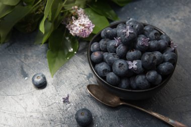 Üzerinde mavi gri bir masada leylak çiçekleri olan yaban mersini yığını manzarası. Antik gümüş kaşık ve leylak dalları vurguluyor..