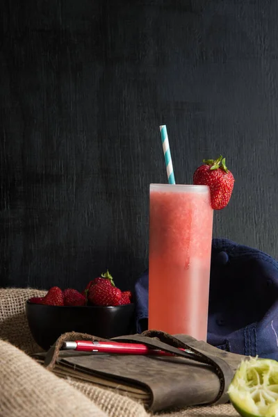 Refreshing, icy strawberry cocktail topped with a strawberry and an eco friendly, festive paper straw, sitting on a table with bowl of fresh strawberries,  a diary,  cap, and squeezed lime.