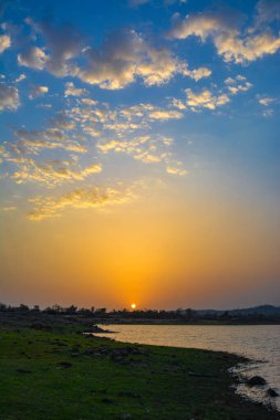 Bir göl ya da nehir üzerinde altın gün batımı canlı mavi gökyüzü ve çimenli bir kıyı şeridinin üzerindeki tüylü bulutlar