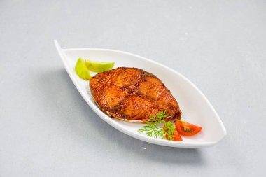 Kahin balık kızartması, soğan, limon ve domates dilimleriyle süslenmiş, beyaz seramik tabakta ve gri desenli arka planda güzelce düzenlenmiş..