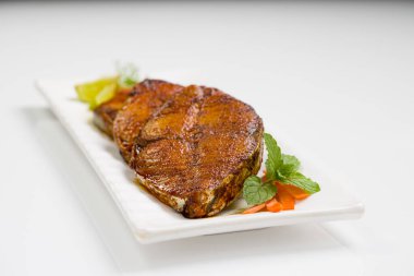 Kahin balık kızartması soğan, limon ve domates dilimleriyle süslenmiş beyaz dikdörtgen şeklinde seramik tabakla beyaz desenli arka plana yerleştirilmiş..