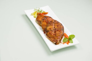 Kahin balık kızartması soğan, limon ve domates dilimleriyle süslenmiş beyaz dikdörtgen şekilli seramik tabakla buz mavisi arka plana yerleştirilmiş..