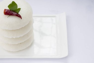 Idly ya da Idli, kırmızı acı biber ve köri yapraklarıyla süslenmiş Güney Hindistan ana kahvaltı öğesi beyaz desenli arka planda beyaz dikdörtgen üzerine güzelce yerleştirilmiş..