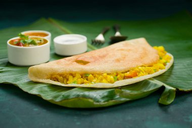 Dosa _ Masala dosa, ünlü Güney Hindistan kahvaltı öğesi geleneksel yöntemle kast demir tavasından yapılır ve koyu yeşil arka planlı, izole edilmiş taze bir muz yaprağı üzerinde düzenlenir..