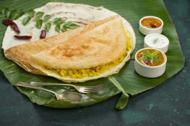 Dosa _ Masala dosa, ünlü Güney Hindistan kahvaltı öğesi geleneksel yöntemle kast demir tavasından yapılır ve koyu yeşil arka planlı, izole edilmiş taze bir muz yaprağı üzerinde düzenlenir..