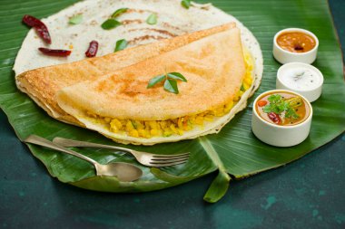 Dosa _ Masala dosa, ünlü Güney Hindistan kahvaltı öğesi geleneksel yöntemle kast demir tavasından yapılır ve koyu yeşil arka planlı, izole edilmiş taze bir muz yaprağı üzerinde düzenlenir..