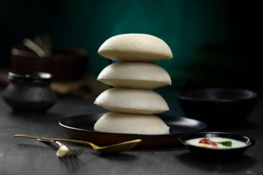 Idly ya da Idli, Güney Hindistan ana kahvaltı öğesi bir yığın üzerine yerleştirilir güzel bir şekilde siyah bir tabakta yerleştirilmiş mutfak arka planı ile koyu yeşil doku, seçici odaklı.
