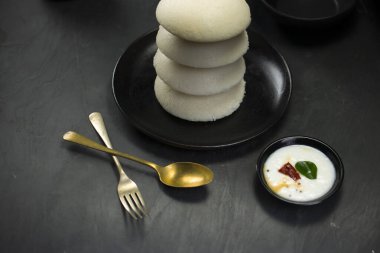 Idly ya da Idli, Güney Hindistan ana kahvaltı öğesi bir yığın üzerine yerleştirilir güzel bir şekilde siyah bir tabakta yerleştirilmiş mutfak arka planı ile koyu yeşil doku, seçici odaklı.