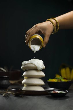 Idly ya da Idli, Güney Hindistan ana kahvaltı öğesi bir yığın üzerine güzel bir şekilde yerleştirilir ve bir kadın boş boş hindistan cevizi turşusu döker koyu yeşil desenli siyah bir tabakta, mutfak arka planında.