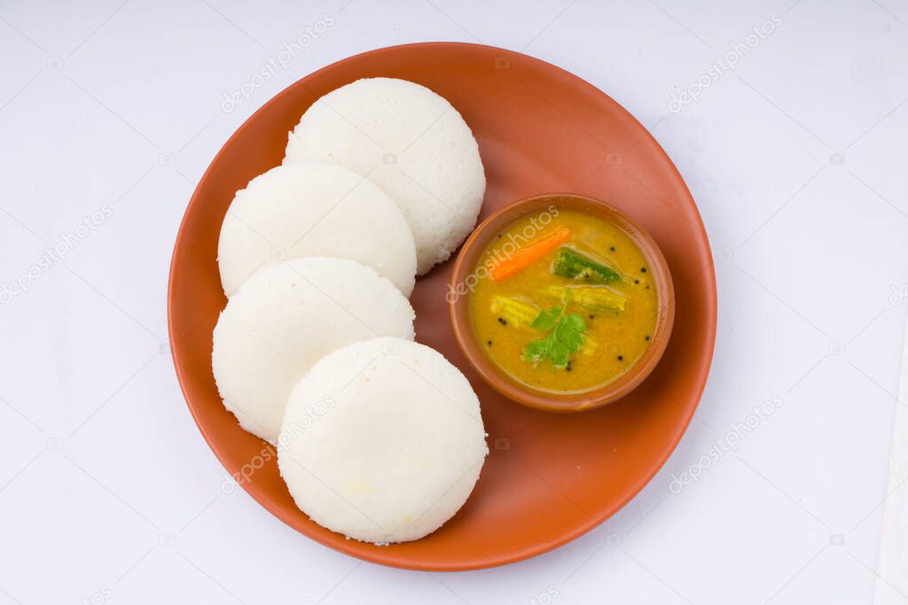 Idly or Idli, south indian main breakfast item which is beautifully arranged in an earthen ware with a small bowl of sambar placed on white background.